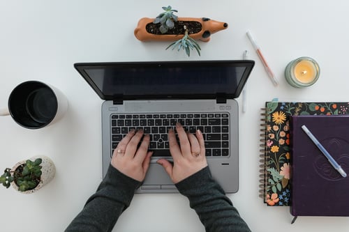 Hands typing at a computer with notebooks, candle, plant, and coffee surrounding workspace