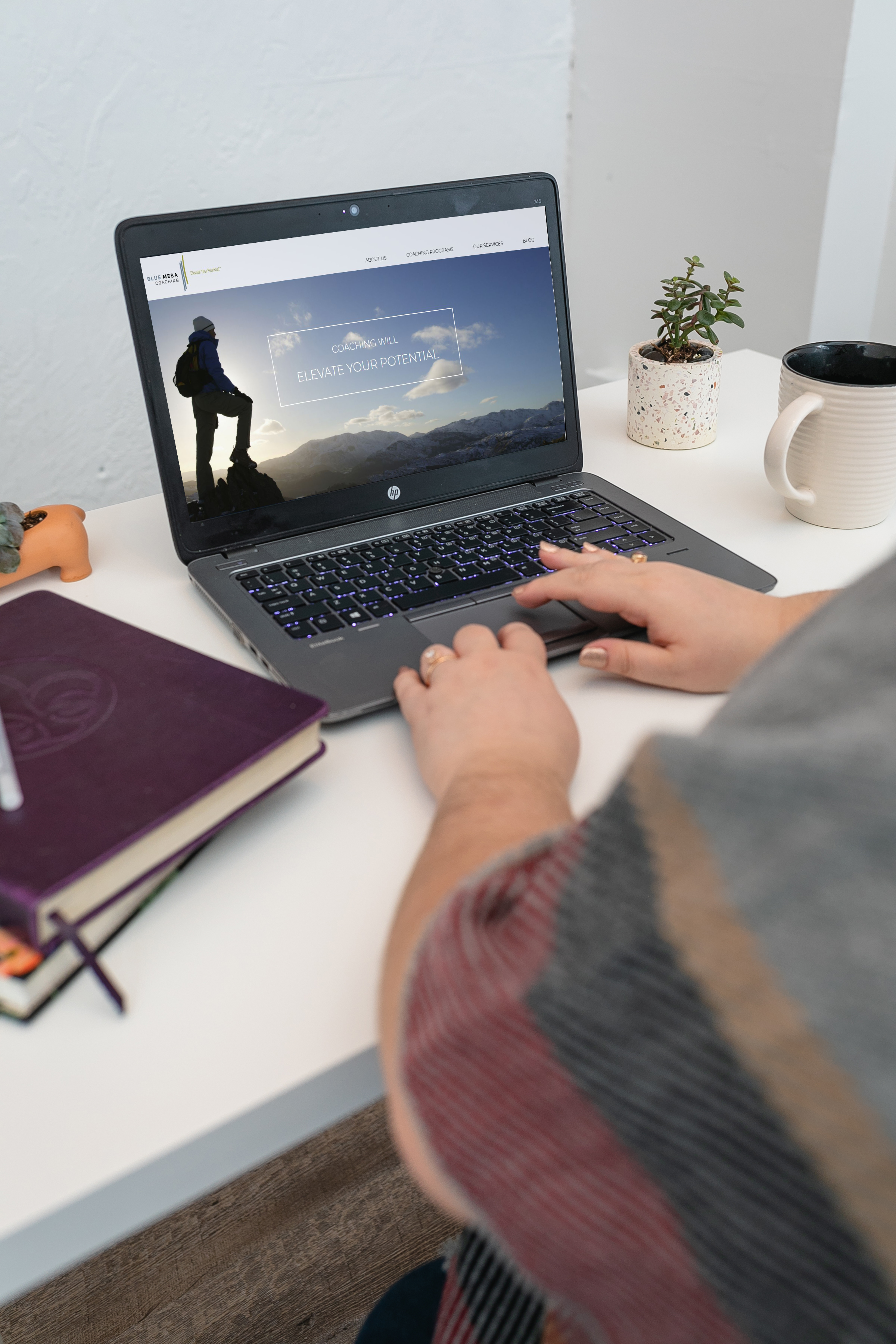 Cut off view of person typing at a laptop with journals and coffee mug surrounding them.