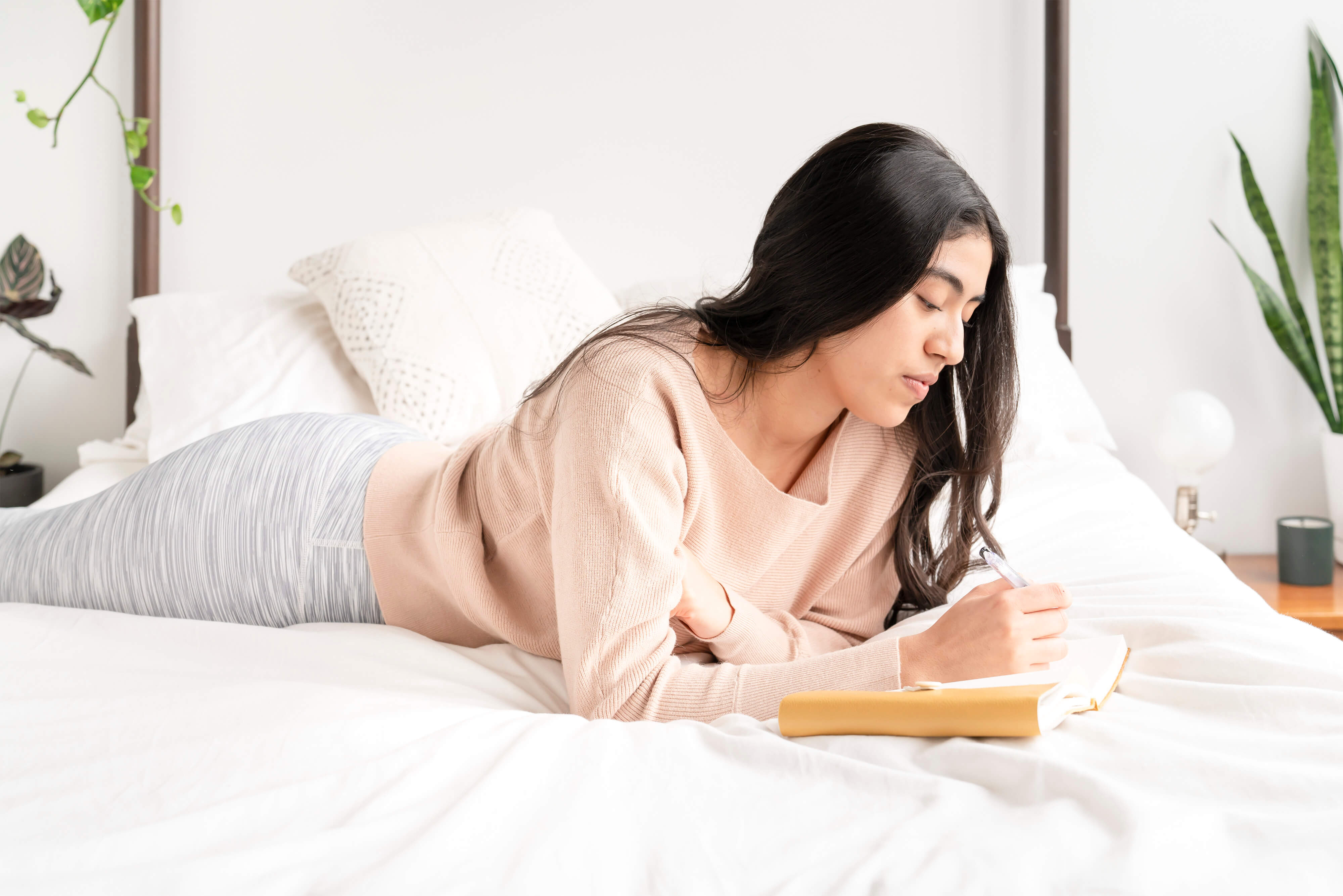 Woman lies on her stomach on a bed while writing in a journal.