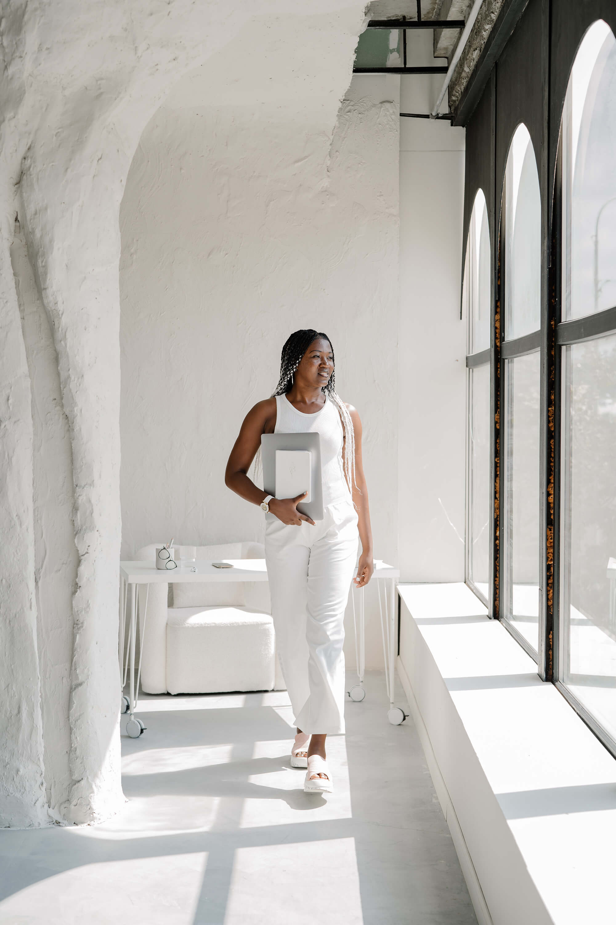 woman in white outfit walks down a window lined hallway holding her computer