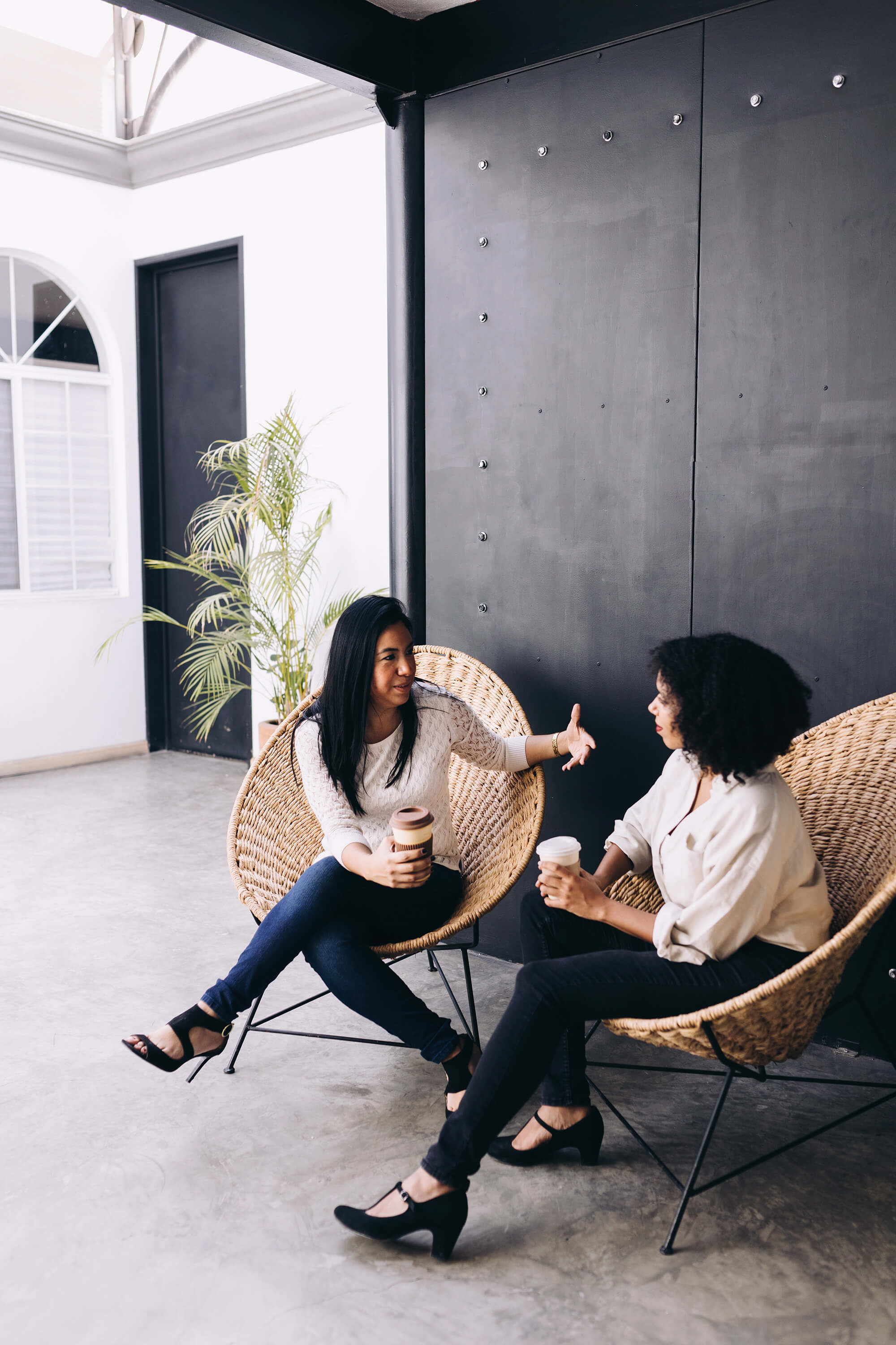 two women sit in egg style chairs holding coffee while having a conversation